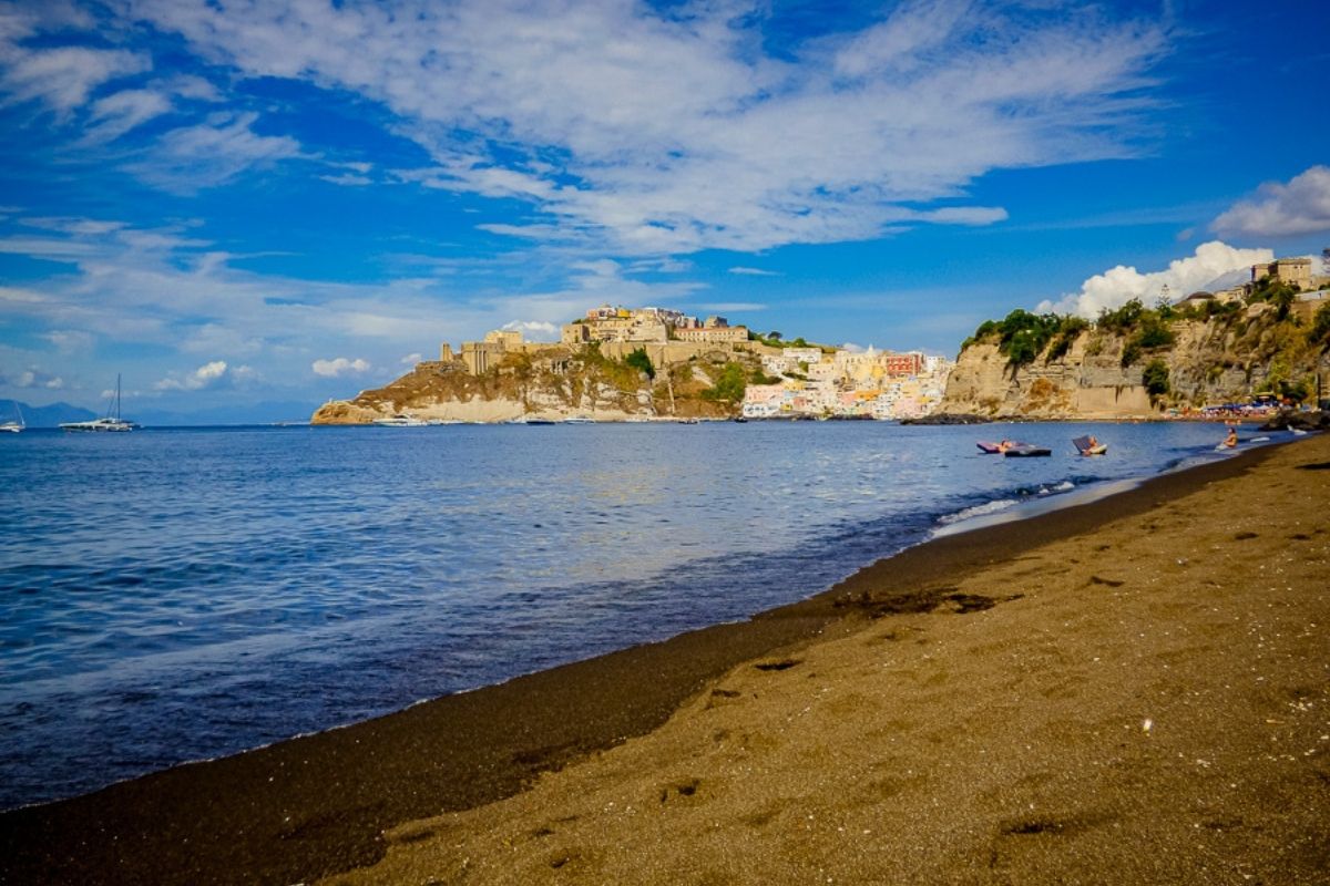 Spiaggia di sabbia vulcanica nera in Italia, con onde che si infrangono e scogli sullo sfondo.